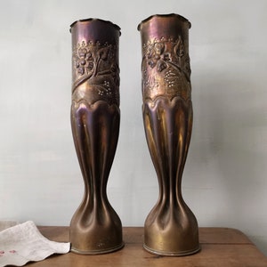 Puede incluir: Dos jarrones antiguos de latón con diseño acanalado y relieve floral. Los jarrones tienen un borde superior con volantes y un acabado metálico cálido. Se exhiben sobre una superficie de madera.