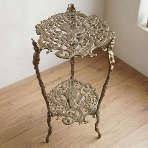 May include: A two-tiered metal plant stand with a brass-toned finish. The stand features intricate, openwork scroll patterns on both the top and bottom circular shelves, supported by three slender, curved legs with decorative detailing.