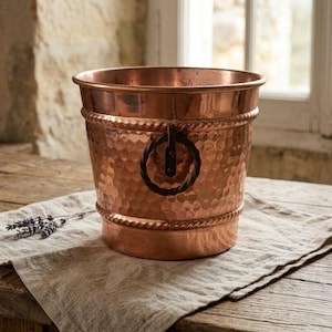 May include: A round, hammered copper bucket featuring a polished metallic finish, a wide top rim, and a decorative rope-style border. The bucket includes a dark, circular metal ring handle attached to the side for a functional, textured appearance.