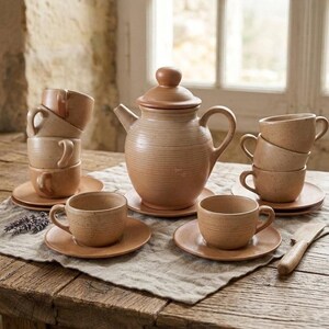 May include: A terracotta tea set featuring a central teapot with a lid and handle, surrounded by several matching cups and saucers. The ceramic pieces have a ribbed, horizontal texture and a natural, earthy tan finish, arranged on a linen cloth.