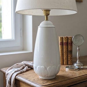 Könnte beinhalten: Eine Tischlampe mit einem glänzenden weißen Keramikfuß, der am unteren Rand ein skulpturales Blütenblattdesign aufweist. Die Lampe hat eine Messingfassung und einen breiten, cremeweißen Stoffschirm. Der Fuß ist glatt und konisch geformt.