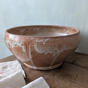 Könnte beinhalten: Eine handgefertigte Keramikschale mit einem breiten, flachen Design. Die Schale hat eine warme, erdige Farbpalette mit Braun- und Weißtönen, die eine rustikale Ästhetik erzeugt. Die Innen- und Außenflächen haben eine strukturierte Oberfläche.