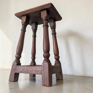 Puede incluir: Un taburete de madera marrón oscuro con una parte superior rectangular y cuatro patas torneadas conectadas por un marco base resistente. El taburete presenta una veta de madera visible y un acabado profundo y rico en toda su estructura.