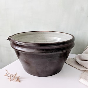 May include: A large, round ceramic mixing bowl with a dark brown, smooth exterior and a contrasting light cream-colored interior. The bowl features a wide, thick rim and a small, integrated pouring spout on the left side, with visible horizontal ridges.