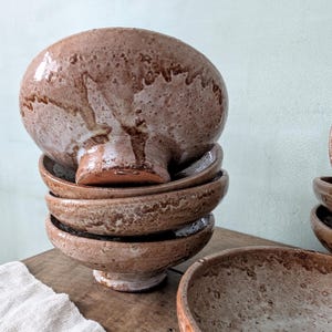 May include: A collection of handmade ceramic bowls with a distinctive, textured glaze in shades of brown and white. The bowls have a rounded shape and a pedestal base, ideal for serving soups or salads. The rustic design adds a touch of elegance to any kitchen.
