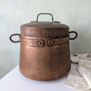 May include: A hammered copper pot with a lid and two side handles. The pot has a riveted band around the middle and a handle on the lid. The copper has a rich, warm color and a textured surface. A vintage kitchenware item.