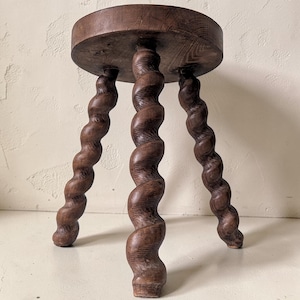May include: A dark brown wooden stool featuring a circular top and three legs carved into a distinct spiral or barley twist design. The wood grain is visible across the surface of the round seat and the three spiraled legs that support the structure.