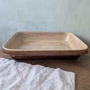 May include: A rectangular, ceramic baking dish with a light brown interior and a darker brown rim. The dish is empty and sits on a wooden surface, with a white cloth in the foreground. The background is a light blue wall.