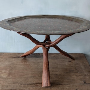 Może przedstawiać: Dekoracyjny stół z okrągłym, ozdobnym metalowym blatem. Taca ma szczegółowy wzór i stoi na drewnianej podstawie wykonanej ze splecionych gałęzi. Unikalny mebel.