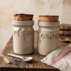 Pode incluir: Dois potes de armazenamento cilíndricos de cerâmica com tampas de cortiça natural. O pote da esquerda apresenta o texto em relevo "sel fin" e o da direita "farine". Ambos os potes têm um acabamento esmaltado em bege claro.
