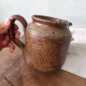 Puede incluir: Una jarra de cerámica marrón con un esmalte moteado y una amplia asa redondeada. La jarra tiene un aspecto rústico y hecho a mano.