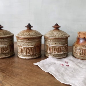 Puede incluir: Un conjunto de cuatro botes de especias de cerámica con tapas y un bote con tapón de corcho. Los botes son de color beige con detalles marrones y llevan las etiquetas "POIVRE", "THYM", "LAURIER" y "CANNELLE".