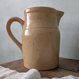 May include: A speckled tan ceramic jug with a curved handle and a pouring spout. The jug has a glossy finish. It is displayed on a wooden surface.