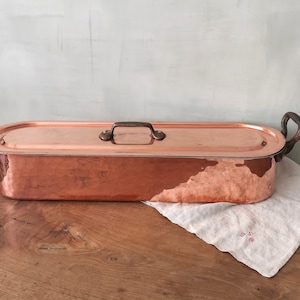 May include: A vintage copper roasting pan with a lid and two handles. The pan is oval shaped and has a hammered finish. It is sitting on a wooden table with a white linen napkin.