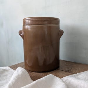 Può includere: Un vaso di ceramica marrone con coperchio, con due manici laterali. Il vaso è cilindrico e ha una finitura lucida. Il coperchio ha delle scanalature orizzontali. Il vaso è appoggiato su una superficie di legno con un panno bianco in primo piano.