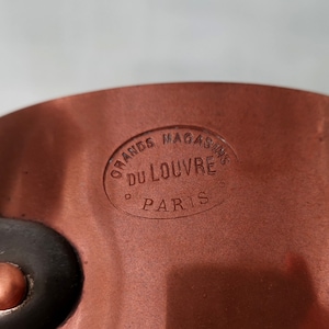 Puede incluir: Primer plano de una olla de color cobre con un asa negra. La olla tiene un sello circular que dice "Grands Magasins Du Louvre Paris".