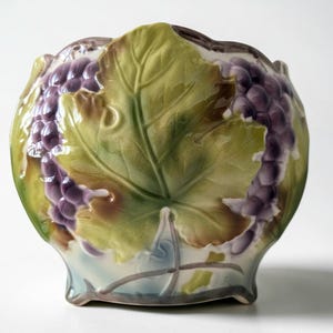 May include: A ceramic bowl featuring a raised, three-dimensional relief design of purple grape clusters and large green vine leaves. The surface has a glossy finish with a scalloped rim and a base that flares slightly outward at the bottom edges.