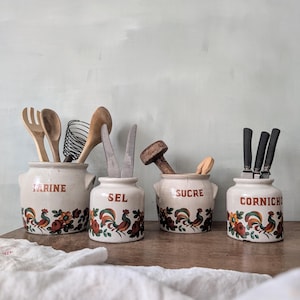 Puede incluir: Cuatro botes de cocina de cerámica con diseños de gallos y flores. Cada bote está etiquetado con una palabra diferente: "FARINE", "SEL", "SUCRE" y "CORNICH". Los botes contienen utensilios de madera y cuchillos.