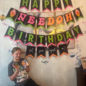 May include: A black, green, and pink banner with the text HAPPY NEEDOH BIRTHDAY and get your squish on. The banner features illustrations of ice cream cones and is displayed on a wall. A person is holding two small boxes of NeeDoh sensory toys.