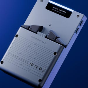 Peut inclure: Une console de jeu portable argent&eacute;e avec des boutons noirs et le texte "BRICK HAMMER". L'appareil a un design &eacute;l&eacute;gant et moderne avec un fond d&eacute;grad&eacute; bleu. Le texte "Better" est visible en bas &agrave; gauche.