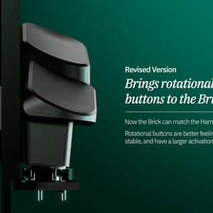 Peut inclure: Un appareil noir avec trois boutons incurv&eacute;s empil&eacute;s, mont&eacute; sur une surface sombre. Le texte "Revised Version Brings rotational buttons to the Brick" est visible. L'appareil a un design moderne et est plac&eacute; sur un fond vert fonc&eacute;.