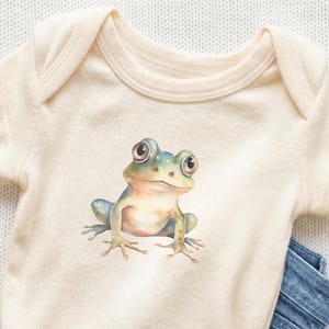 May include: Cream-colored baby onesie featuring a watercolor illustration of a frog. The frog is light blue and green with large, round eyes. The onesie is paired with blue denim jeans, creating a casual outfit for a baby.