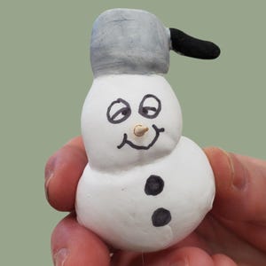 May include: A small, white snowman figurine with a gray hat and black coal buttons. The snowman has a carrot nose and a smiling face. The figurine is held in a person's hand.