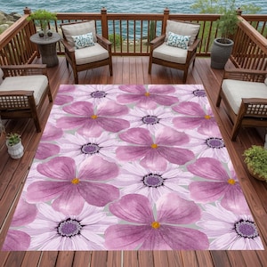 Puede incluir: Una alfombra grande para exteriores con un patrón floral repetido de flores moradas y blancas con centros amarillos. La alfombra está colocada en una terraza de madera con sillas y vistas al agua.