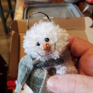 May include: A small, white snowman ornament with a fluffy texture. It has black bead eyes, a carrot nose, and a stitched smile. The snowman wears a green plaid scarf and has a metal loop for hanging. A button is sewn on the front.