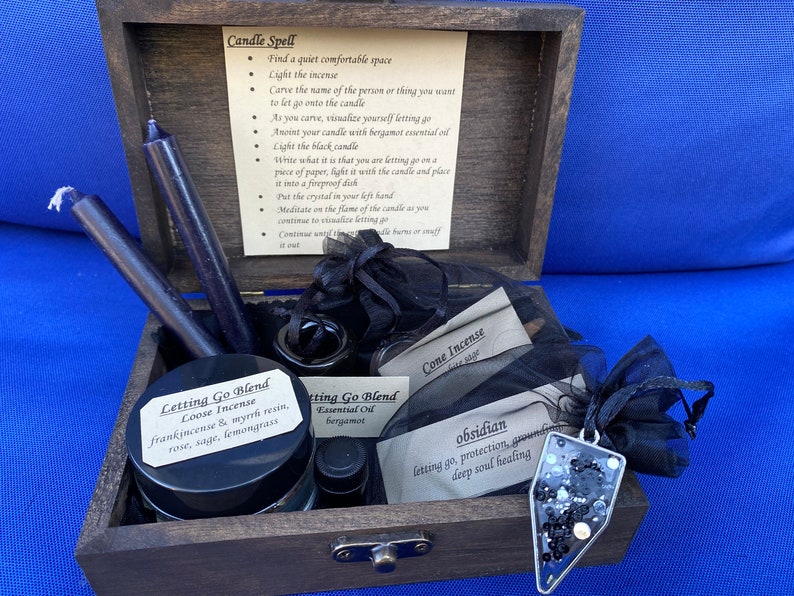 May include: A wooden box set containing ritual items. Inside are two dark purple candles, a jar of loose incense, essential oil, cone incense, and an obsidian pendant. The box also includes instructions for a candle spell.