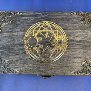 May include: A rectangular wooden box with a gray finish and ornate gold-colored metal corner accents. The box features a gold circular emblem with celestial and geometric designs on the lid. The box is closed and sits on a blue surface.