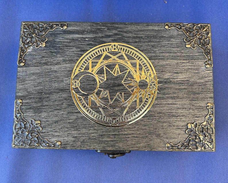 May include: A rectangular wooden box with a gray finish and ornate gold-colored metal corner accents. The box features a gold circular emblem with celestial and geometric designs on the lid. The box is closed and sits on a blue surface.