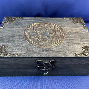 May include: A rectangular wooden box with a gray finish, adorned with ornate brass corner accents and a central circular design. The box features a metal clasp and hinges, suggesting a storage or keepsake purpose.