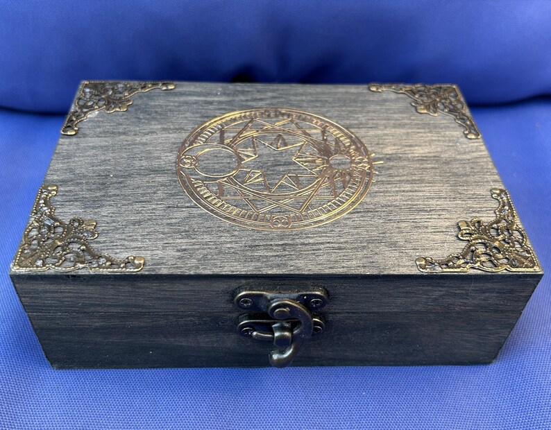 May include: A rectangular wooden box with a gray finish, adorned with ornate brass corner accents and a central circular design. The box features a metal clasp and hinges, suggesting a storage or keepsake purpose.