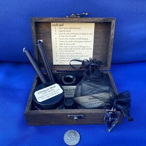 May include: A wooden box set containing black candles, incense, essential oils, a crystal pendant, and instructions for a candle spell. The set includes a jar labeled "Letting Go Blend" and a small bag with the word "Obsidian" on it.