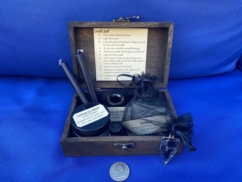 May include: A wooden box set containing black candles, incense, essential oils, a crystal pendant, and instructions for a candle spell. The set includes a jar labeled "Letting Go Blend" and a small bag with the word "Obsidian" on it.
