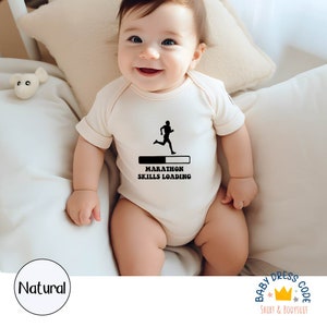 Könnte beinhalten: Ein weißer Baby-Strampler mit einer schwarzen Grafik einer laufenden Person und dem Text "Marathon Skills Loading".