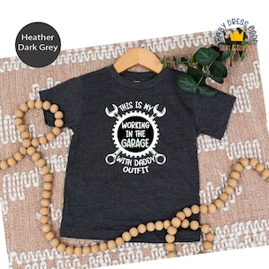 Op de afbeelding: Donkergrijs gemêleerd T-shirt met korte mouwen met een witte afbeelding van gekruiste schroevendraaiers en een tandwiel met de tekst "This is my working in the garage with daddy outfit".