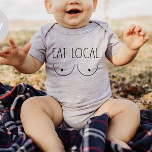 Puede incluir: Un bebé con un body gris que dice "EAT LOCAL" y una imagen estilizada de pechos. El bebé está sentado en una manta a cuadros.