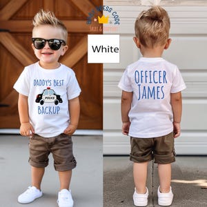 May include: Two children wearing white t-shirts with blue text. The shirt on the left says "Daddy's Best Backup" with a cartoon police car. The shirt on the right says "Officer James". Both children are wearing khaki shorts and white sneakers.