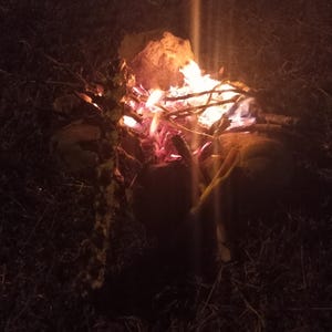 May include: A campfire at night, with flames of orange and red. The fire is surrounded by rocks and small branches. The scene is dimly lit, with a vertical streak of light.