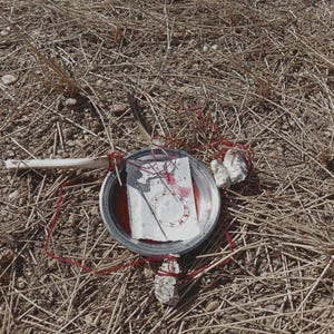 Może przedstawiać: Scena rytualna z metalową miską z czerwoną cieczą i papierem z okrągłymi oznaczeniami. Misa połączona jest czerwoną nicią z dwoma kamieniami i drewnianym patykiem, wszystko na suchej trawie i słomie.