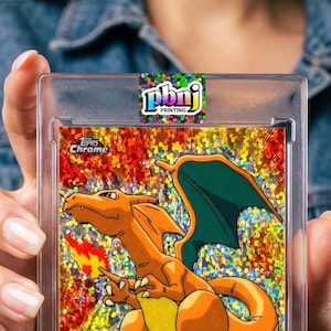 May include: A Topps Chrome Charizard trading card encased in a clear protective plastic holder. The card features a holographic orange dragon-like creature with green wings, a yellow belly, and a flame tail against a shimmering gold background.