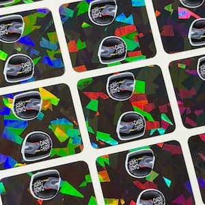 May include: A sheet of holographic stickers with a black background and colourful, geometric shapes. Each sticker features a white and black design with the text "DADS CARDS".