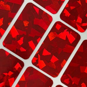May include: Close-up of several rectangular stickers with a vibrant red, glittery finish. Each sticker is filled with irregular, reflective shapes, creating a dazzling effect. The stickers are arranged on a white background.