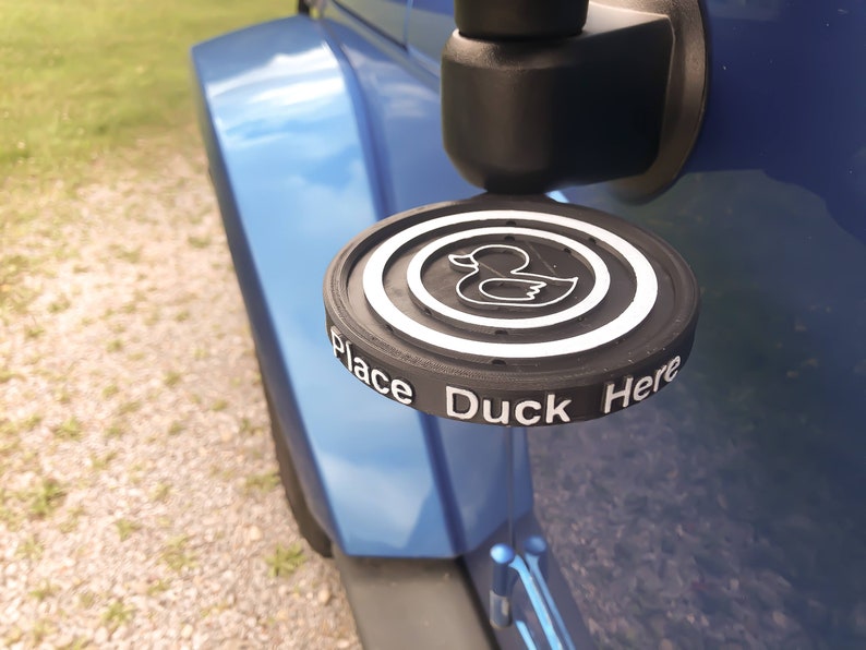 May include: A black and white rubber ducky holder with the words "Place Duck Here" around the edge. The holder is attached to a blue car.