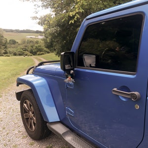 May include: A blue Jeep Wrangler with a black side mirror and a small toy figure attached to the side door handle. The Jeep is parked on a gravel driveway with a grassy field in the background.