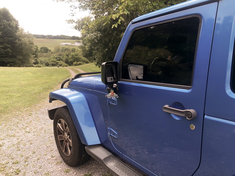 May include: A blue Jeep Wrangler with a black side mirror and a small toy figure attached to the side door handle. The Jeep is parked on a gravel driveway with a grassy field in the background.