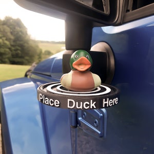 May include: A black plastic duck holder attached to a blue car. The holder has the words "Place Duck Here" printed on it. A rubber duck with a green head, brown body, and white accents is sitting on the holder.