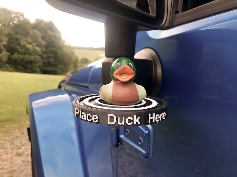 May include: A black plastic duck holder attached to a blue car. The holder has the words "Place Duck Here" printed on it. A rubber duck with a green head, brown body, and white accents is sitting on the holder.